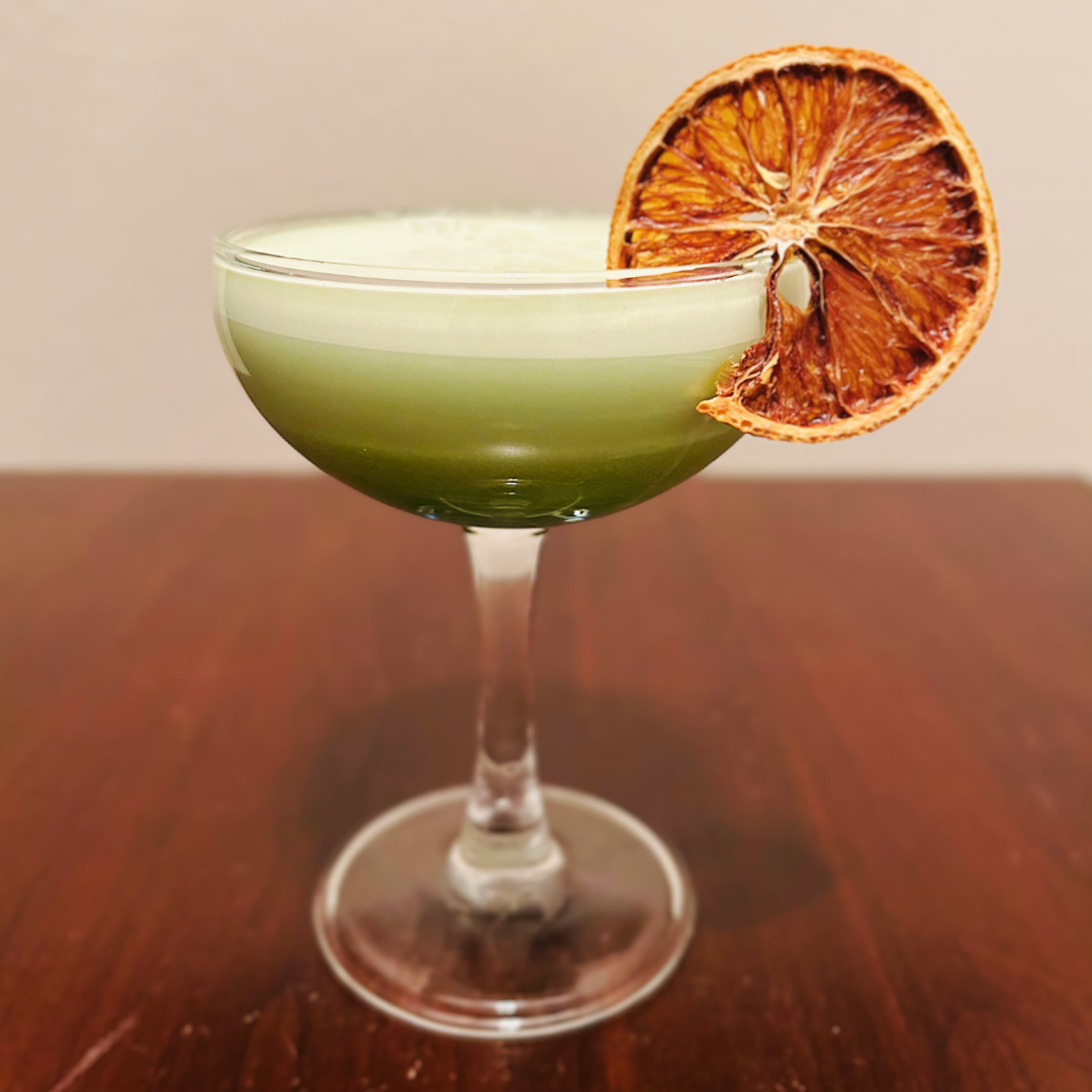 coupe glass with dark green matcha cocktail on bottom and frothy white foam on top. Garnished with orange wheel