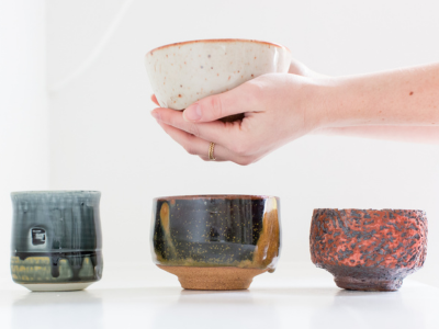 hands holding a off white tea bowl. Underneath are different tea bowls in blue brown and red