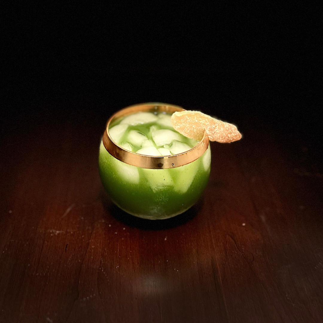 Matcha cocktail in a low ball glass on ice garnished with a slice of crystalized ginger