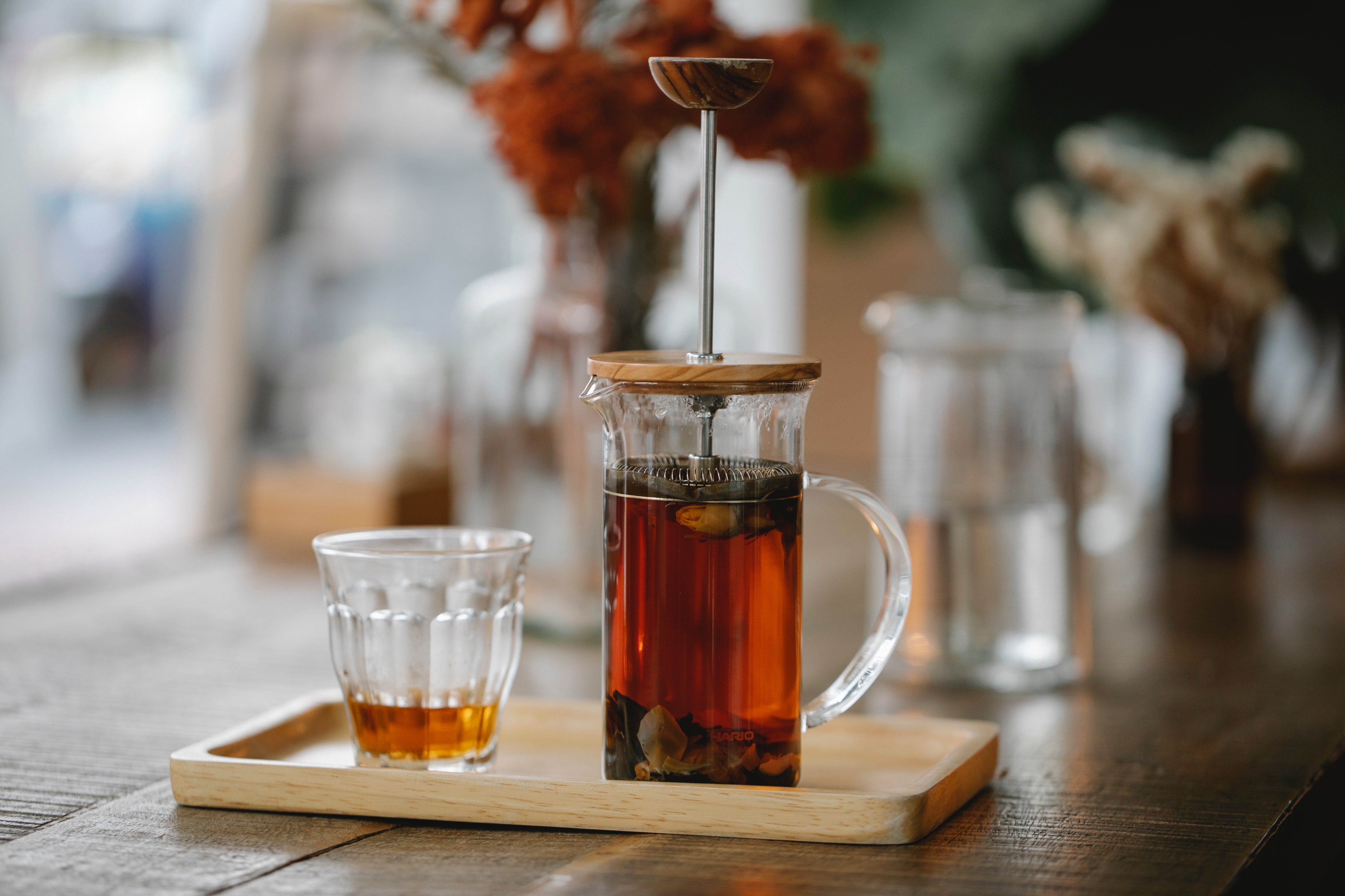 glass french press teapot steeping tea, on wood tray next to glass cup with tea on a restaurant table