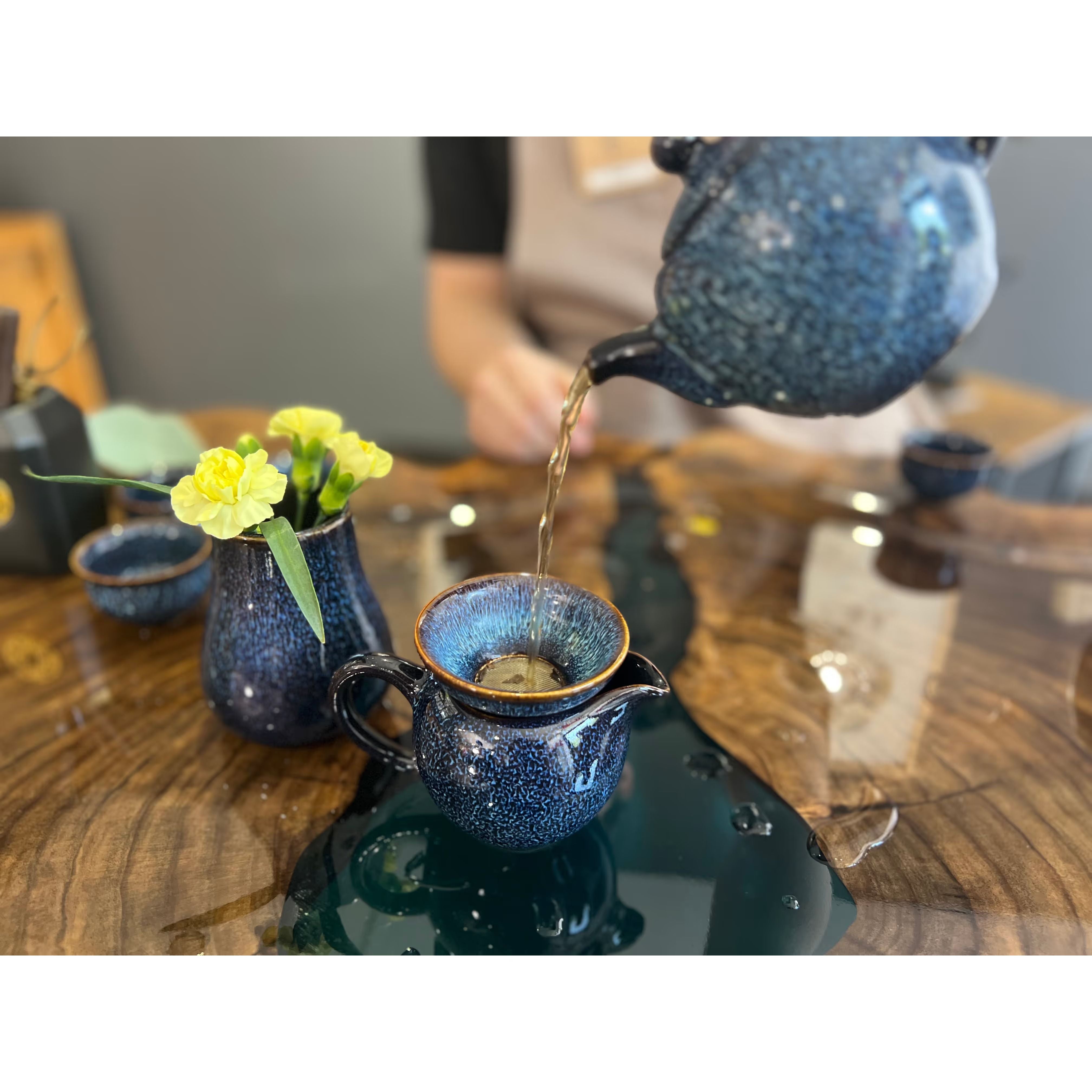 blue glazed sencai teapot pouring artisan tea into matching fair cup next to yellow flowers in matching vase on artisan wood and blue resin tea table with water droplets
